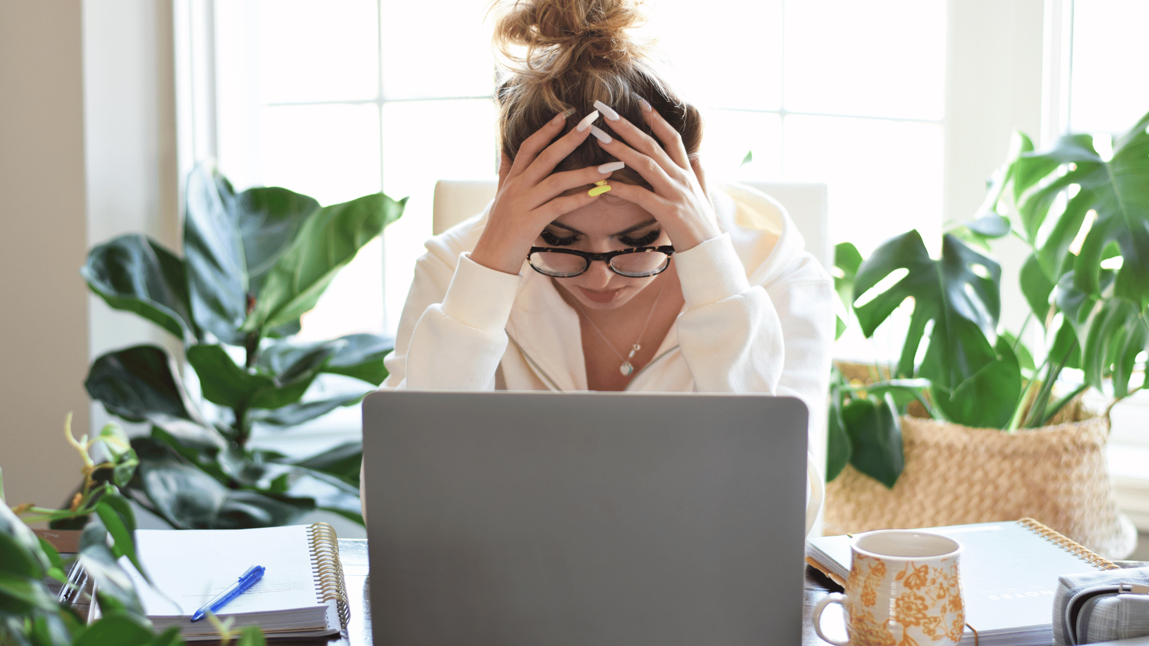 A woman, overwhelmed by her inner critic dialogue, holds her head in her hands while sitting at her work desk, a worried expression on her face and a laptop open in front of her.